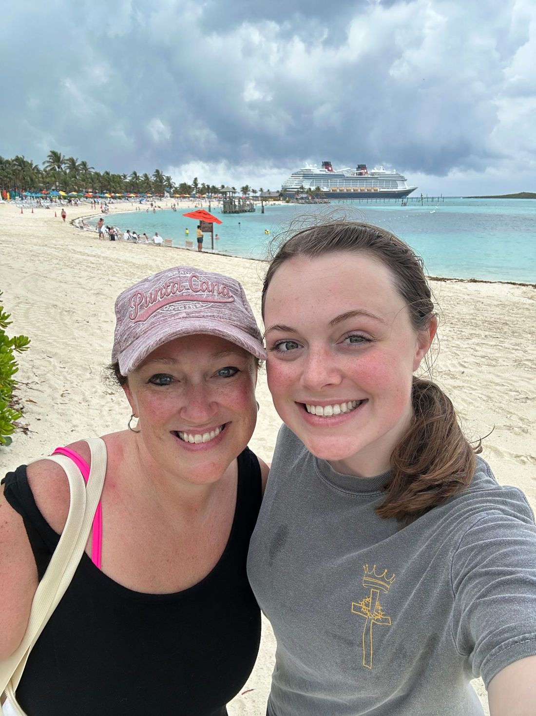 ’Chelle and her daughter smiling on a white-sand beach, with their cruise ship anchored off shore behind them